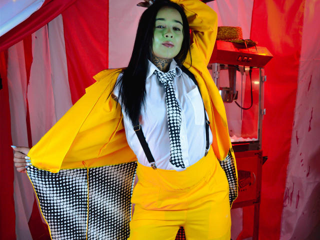BlackRousse with long black hair, green face paint, yellow suit, white shirt, black-and-white tie, and suspenders, standing with arm raised, wearing a confident, sexy, cosplay look.