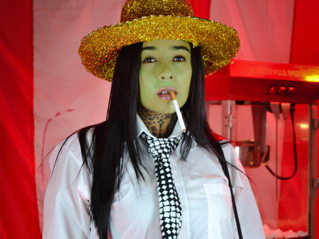 BlackRousse with long black hair, green face paint, gold glitter hat, white shirt, black and white tie, and cigarette in mouth, showing tattoos on neck. Red background.
