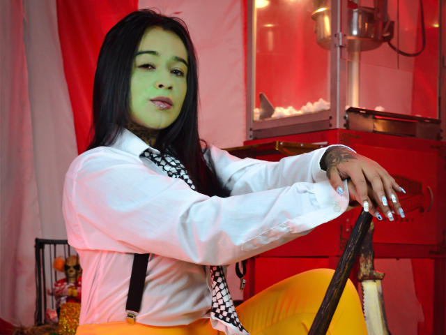 BlackRousse with long black hair, green face paint, white shirt, checkered necktie, and orange pants, leaning on a rusty bike, showing tattooed wrist and silver nails.