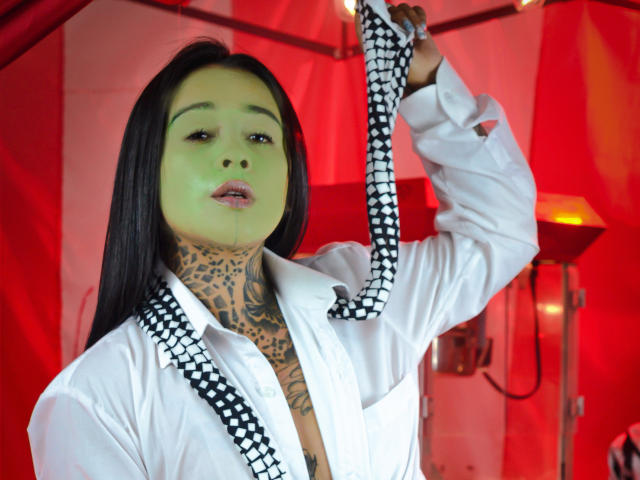 BlackRousse with long black hair, green face, white shirt, black-and-white checkered tie, tattooed neck, holding tie, red-lit background, looking at camera, slightly open shirt.