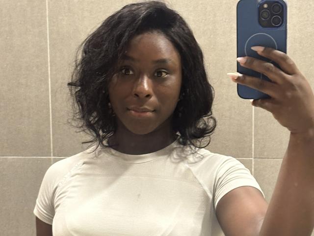 LizaPatrick, à la peau foncée et aux cheveux noirs ondulés, porte un t-shirt blanc et tient un téléphone bleu dans une photo de profil prise dans un miroir, affichant un léger sourire et une silhouette attrayante..