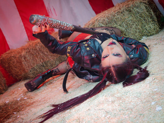 AriannaSeete, with long dark pigtails, lies on hay in a black leather jacket, fishnet stockings, and metallic boots, her red lipstick contrasting her pale skin. She's flipping her middle finger.