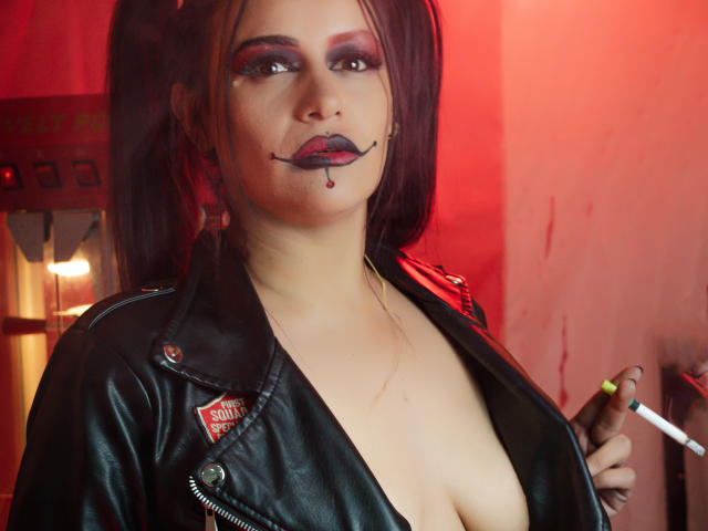 AriannaSeete with black leather jacket, red eye makeup, and black lipstick, holding a cigarette, showing cleavage, dark pigtails, red light, and "Suicide" patch on jacket.