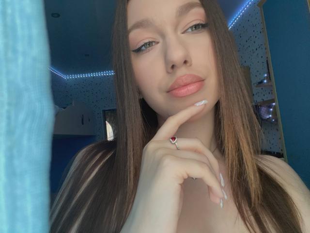 ElveraSatterfield has long brown hair, pink lipstick, and is touching her chin with a finger adorned with a red heart ring, showing off her white nails.