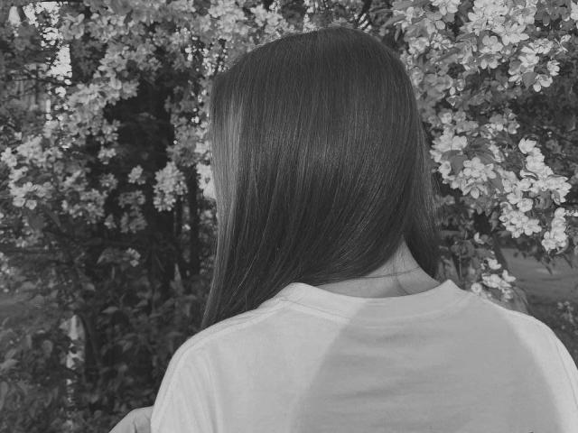 ElveraSatterfield with long, straight hair, wearing a white shirt, standing with her back to the camera, facing a blooming tree filled with small, white flowers. Black-and-white photo.