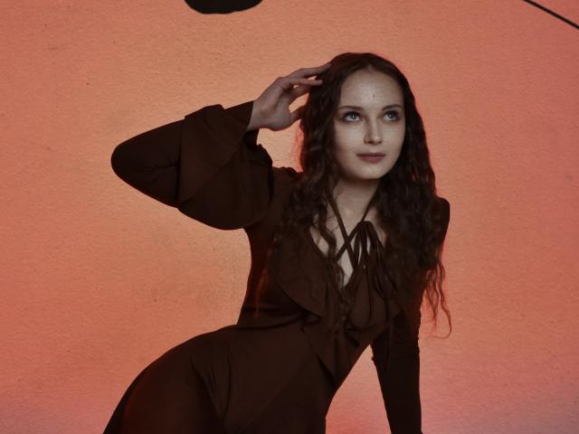 MilenaSparkle with wavy brown hair, wearing a brown dress, has one hand in her hair, looking to the side, standing against a pink-orange wall. Her tits are small.