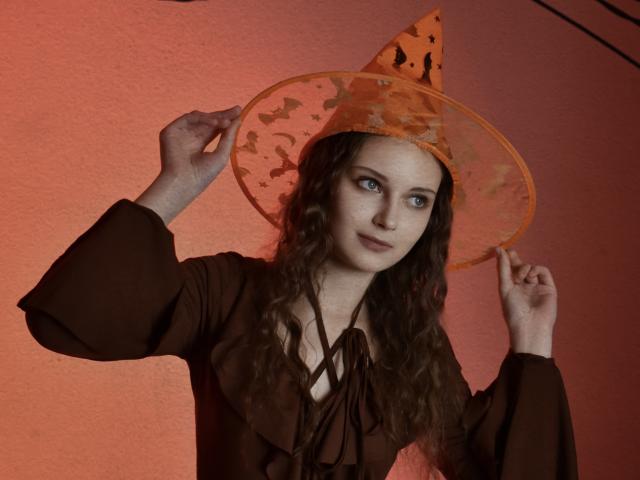 MilenaSparkle with curly brown hair, wearing an orange bat-patterned witch hat, brown dress with ruffled neckline, holding hat, red light background, blue eyes, slight smile, hands up.