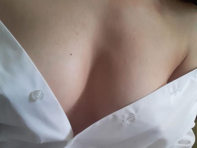 Orshiti's bare chest with a white shirt partially open, revealing a soft, pale skin tone and a slight shadow under the shirt. Boobs visible, buttoned shirt, close-up.