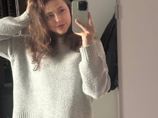 MaidaCain with wavy brown hair, wearing a grey, fuzzy sweater, taking a mirror selfie with a hand in her hair, holding a silver phone, smiling slightly.