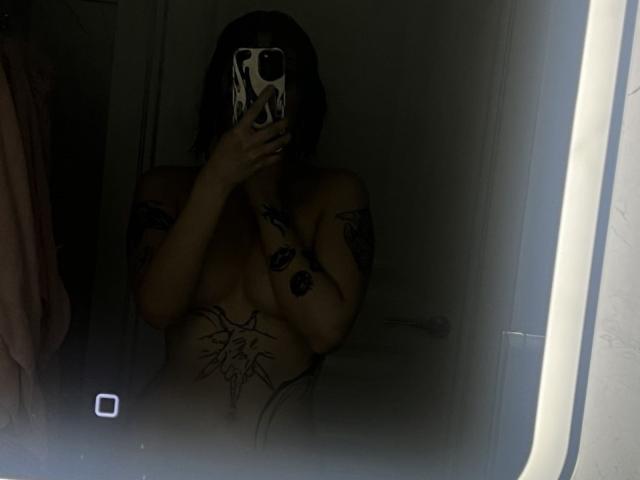 Nude WenMewborn with tattoos, dark hair, holding phone covering face, small tits, dark room, mirror reflection, phone case with white pattern, dim light, towel partially visible.