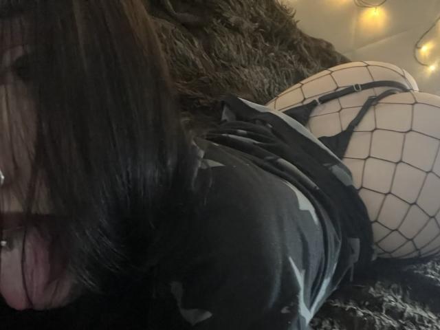 WenMewborn with black fishnet stockings, black thong, and black top, lying on a dark furry surface, showing her ass and a nose ring, with dark hair in the foreground.