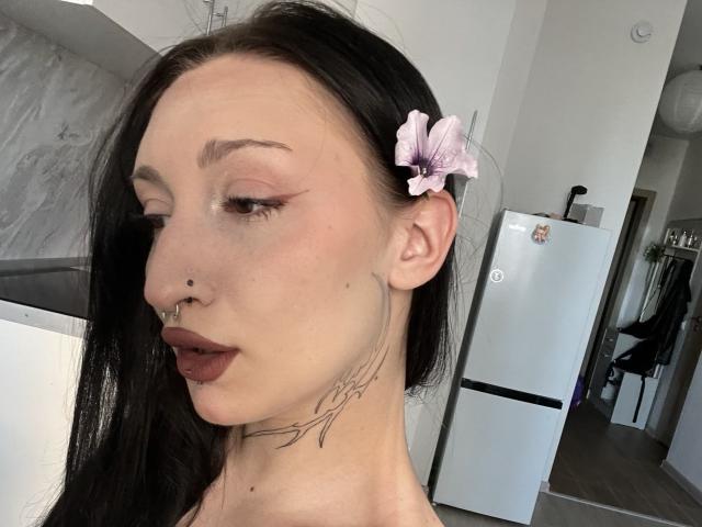 WenMewborn with long black hair, pink flower in hair, nose ring, lip ring, maroon lipstick, and a delicate tattoo on the neck. White refrigerator in the background.