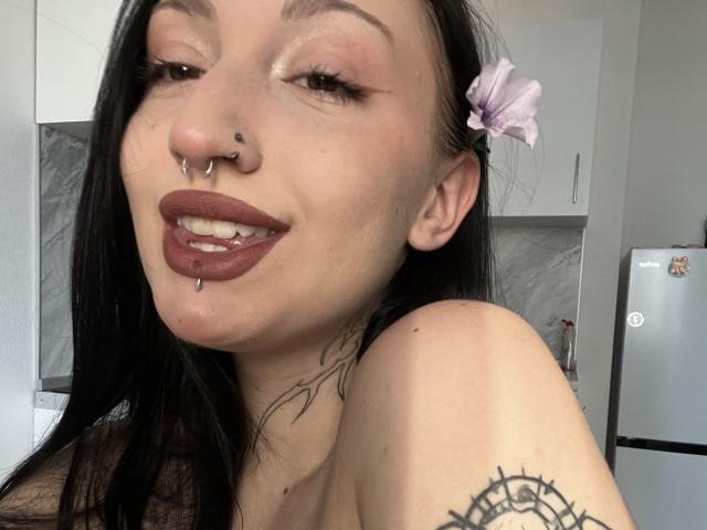WenMewborn has dark hair, a pink flower, nose rings, and lip piercings, with tattoos on her neck and arm, showing off her brown lipstick and smiling.