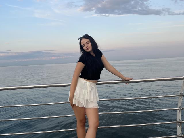 RoxyVesper with long black hair, black top, and white skirt, leaning on a metal railing, ass slightly visible, over calm ocean, sunset sky, sunglasses on head.