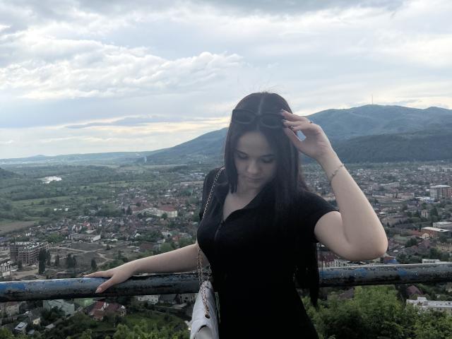 RoxyVesper with big tits, black dress, and long dark hair, stands against a railing, holding sunglasses, looking down, mountains and city in the cloudy background, fair-skinned.