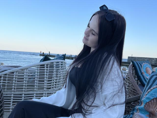 RoxyVesper with long black hair, wearing a white shirt and black top, sitting on wicker chairs, sunglasses on head, looking sideways, blue sky, ocean, and patterned pillows in background.