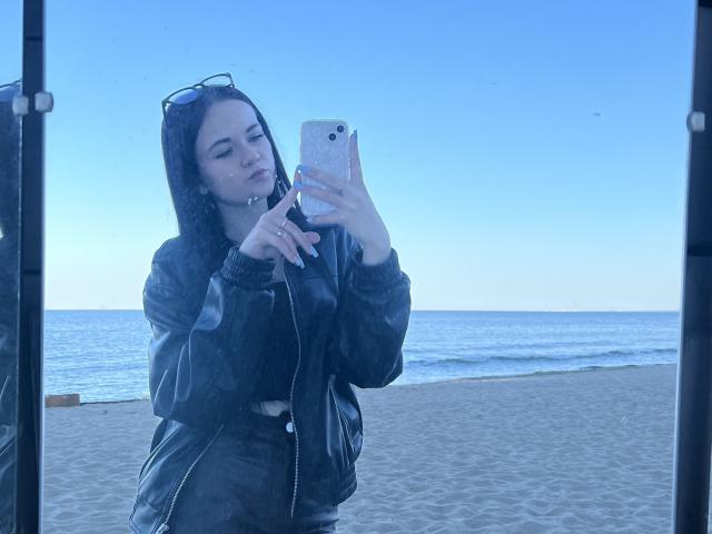 RoxyVesper with dark hair, black leather jacket, and black pants, taking a selfie in a mirror on a beach, holding a phone with a pink case. Blue sky, ocean.