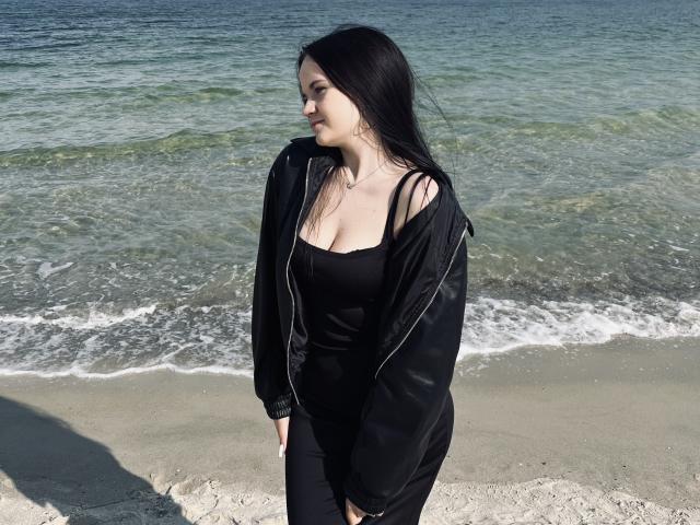 RoxyVesper with black hair, black dress, and open black jacket standing on a beach, showing small tits and looking sideways, ocean and sand in background.