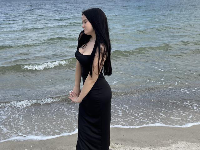RoxyVesper stands on a beach in a black, form-fitting dress, her long black hair flowing, looking sideways with hands clasped. She has a curvy, sexy ass and small tits.
