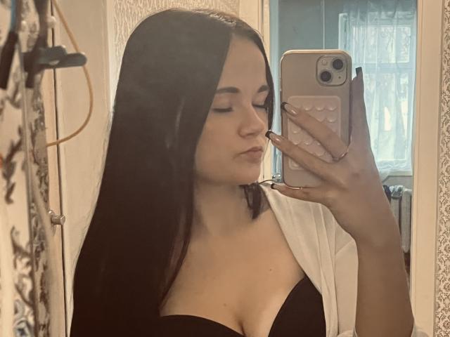 RoxyVesper with long black hair, pouty lips, wearing a black bra and white robe, taking a mirror selfie with her phone, showing her tits in the reflection.