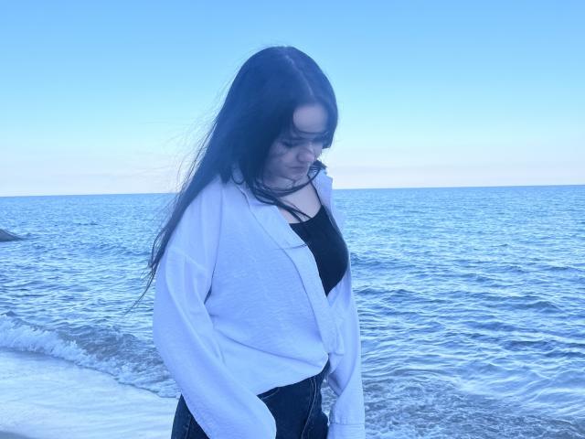 RoxyVesper with long dark hair, wearing a white sweater over a black top, standing by the blue ocean, looking down with a slight smile, hands in her jeans.