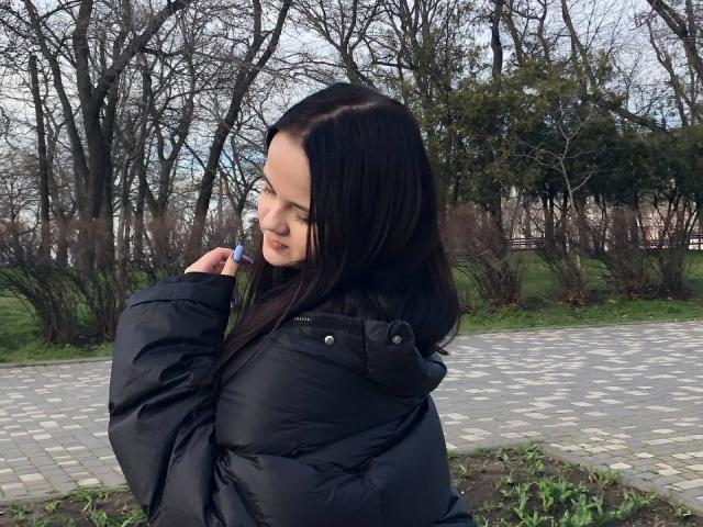 RoxyVesper with dark brown hair, wearing a black puffy jacket, holding a green leaf, looking sideways, in a park with a gray brick path and bare trees.