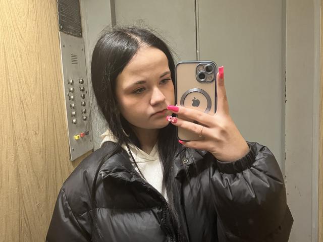 RoxyVesper, with black pigtails, pink nails, and a puffy black jacket, takes a selfie in an elevator using an iPhone. Her white hoodie is partially visible.