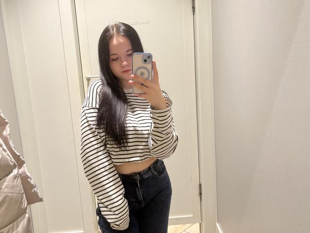 RoxyVesper with long black hair, striped crop top, and high-waisted jeans, taking a mirror selfie in a store. She's holding a phone with a blue case.