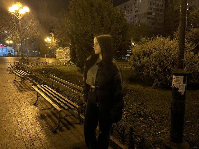 RoxyVesper with long brown hair, wearing a black coat and blue shirt, standing on a brick path at night, next to metal benches, under warm streetlights.