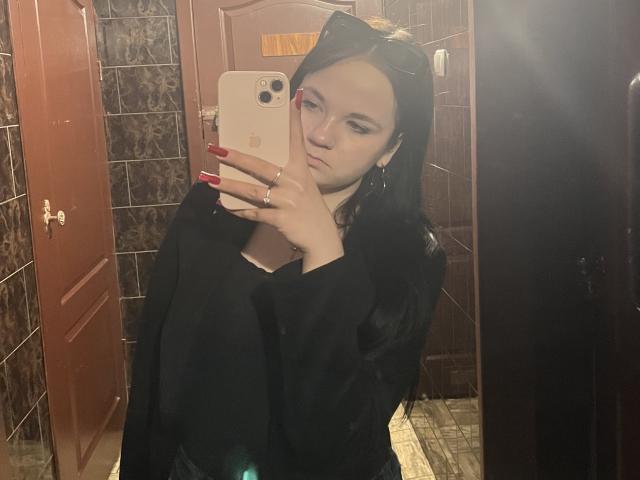 RoxyVesper with long black hair, cat ears, red nails, black top, holding iPhone, mirror selfie, looking serious, light skin, brown tiled bathroom, wooden door.