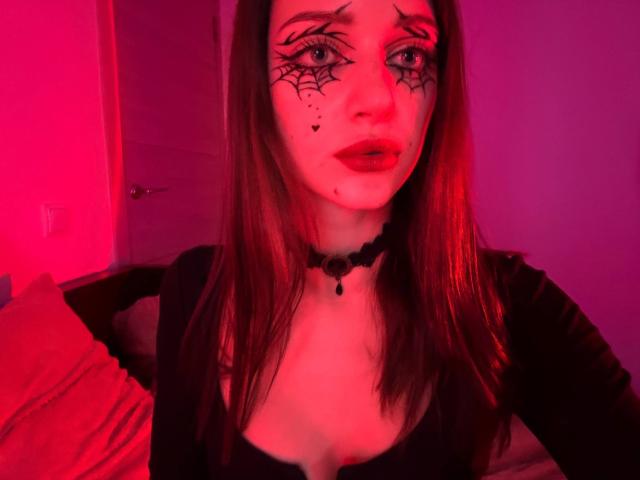 JoelleCalixte with red-pink lighting, black spider web makeup, black choker, black top, showing cleavage, red pillow, red lipstick, brown hair, and a slightly sad expression.