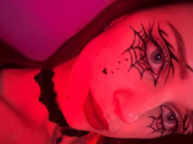 JoelleCalixte with red-lit face, spiderweb makeup on eyes, black choker, and dark red lipstick, staring intensely at camera. Her blue eyes and long brown hair are prominent.