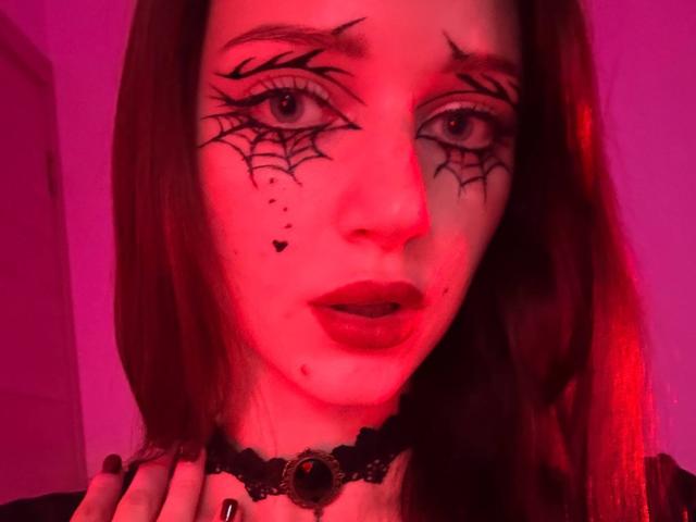 JoelleCalixte's face is red-lit with spiderweb makeup on her eyes, dark lipstick, and a black choker with a gem. She has hand on her neck, looking at camera.