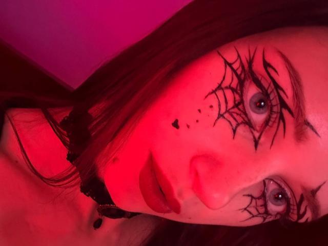 JoelleCalixte's face is red-lit, showing black spiderweb makeup on her eyes, with blue eyes and dark red lipstick. Her black hair is down, and she's wearing a black choker.