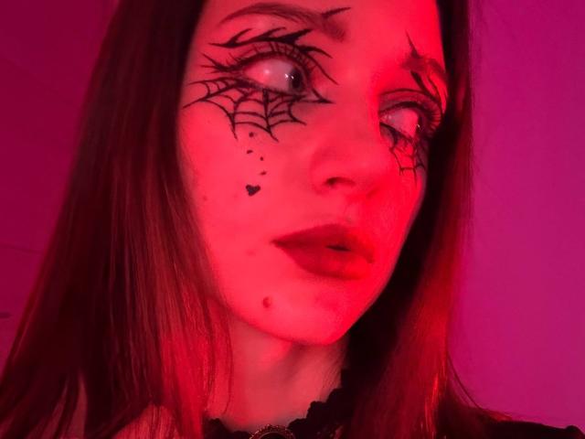 JoelleCalixte with red-tinged face, web tattoos on her eyes, heart and dot tattoos below, and dark lipstick, looking sideways with slightly parted lips. Her brown hair is straight.