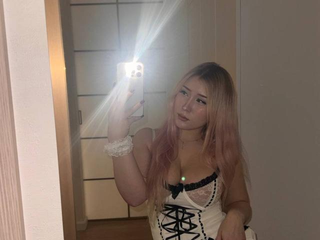 DelphiaCabos with pink hair, wearing a white lace-up corset, mirror selfie, phone flash, white scrunchie, pale skin, small green light on chest, holding phone, looking at camera.