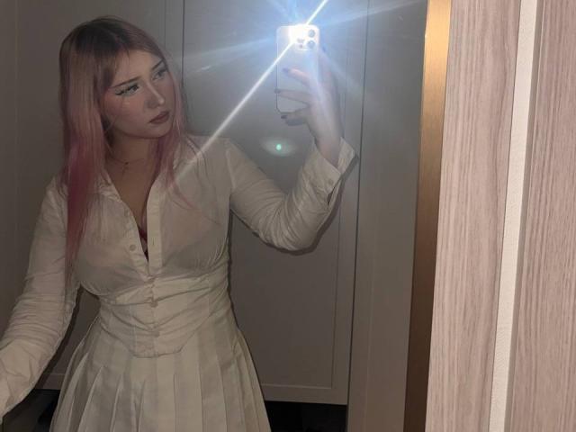 DelphiaCabos with pink hair, wearing a white, button-up, long-sleeve dress, taking a mirror selfie, light from phone flash, slight pout, looking at camera, reflection visible.