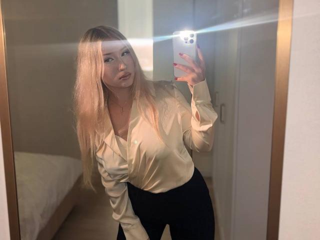 DelphiaCabos with long blonde hair, white satin blouse, and black pants, taking a mirror selfie with a glowing iPhone, showing off her ass and red manicured nails.
