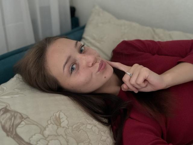 JoelleCalixte with long brown hair, freckles, and blue eyes, lying on a bed, wearing a red shirt, touching her chin with a finger, wearing a ring.