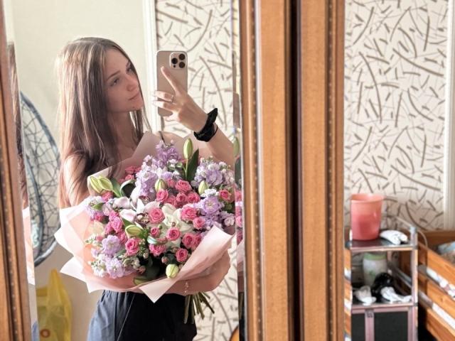 JoelleCalixte with long brown hair, holding a pink and purple flower bouquet in front of a mirror, wearing a black dress, taking a selfie with an iPhone.