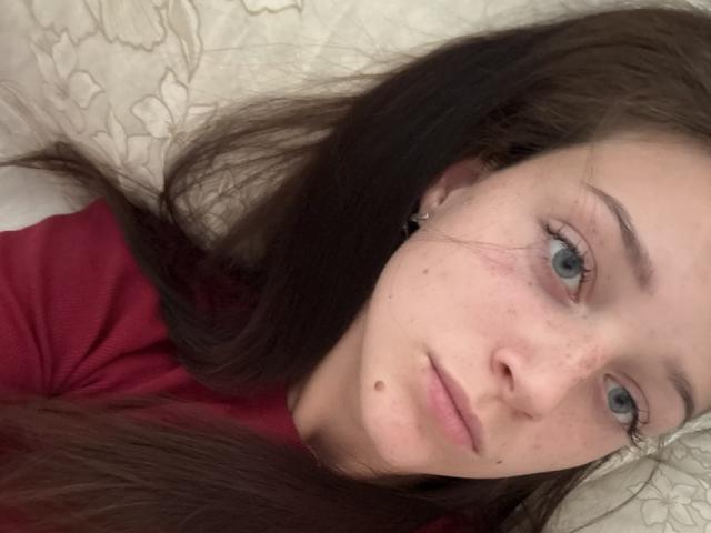 JoelleCalixte has pale skin, blue eyes, long brown hair, small mole on her left cheek, and is wearing a red shirt, lying on a white pillow.