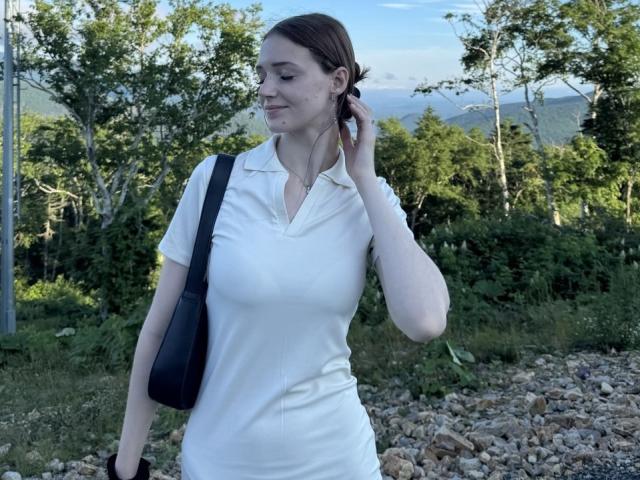 JoelleCalixte with brown hair, white polo shirt, black shoulder bag, holding phone to ear, standing on rocky path, green trees in background, slight smile, looking to the side.