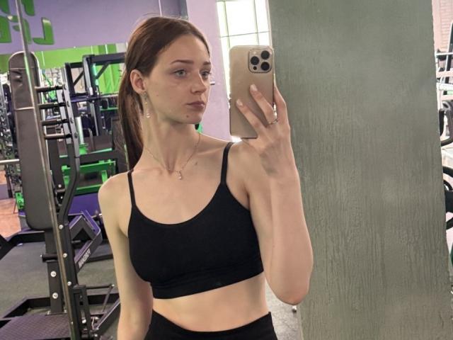 JoelleCalixte with pale skin, brown hair in a ponytail, wearing a black sports bra, holding a phone in a gym, showing a pouty face, small tits, and a slim build.