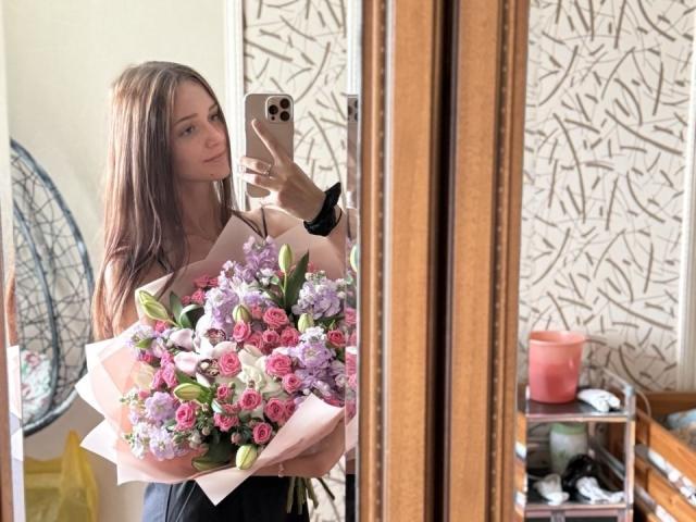 JoelleCalixte with long brown hair, holding a pink bouquet of roses and lilacs, taking a mirror selfie with an iPhone, wearing a black top, in a cozy room.