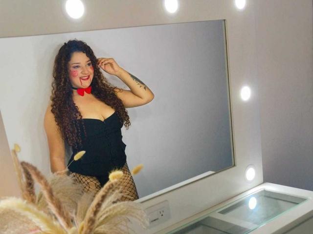 Natalia, mit lockigem, braunem Haar, rotem Fliege, schwarzem Top ohne Träger, Fischernetzstrümpfen, lächelt in den Spiegel, eine Hand im Haar, Spiegelbild, weiße Wände, runde Spiegelbeleuchtung und eine dicke Pflanze im Vordergrund..