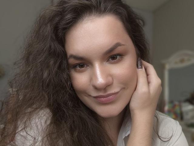 Dreia a les cheveux bruns foncés ondulés, un visage éclatant et des yeux bruns. Elle sourit légèrement, touche sa joue avec une main, porte une chemise blanche et a des ongles brillants..