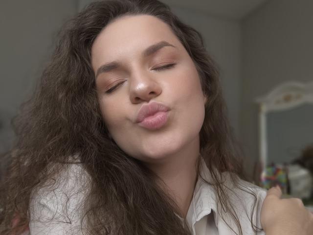 Dreia with puffy pink lips, closed eyes, and wavy brown hair, wearing a white shirt, making a pouty face, holding her own arm, looking cute and relaxed.