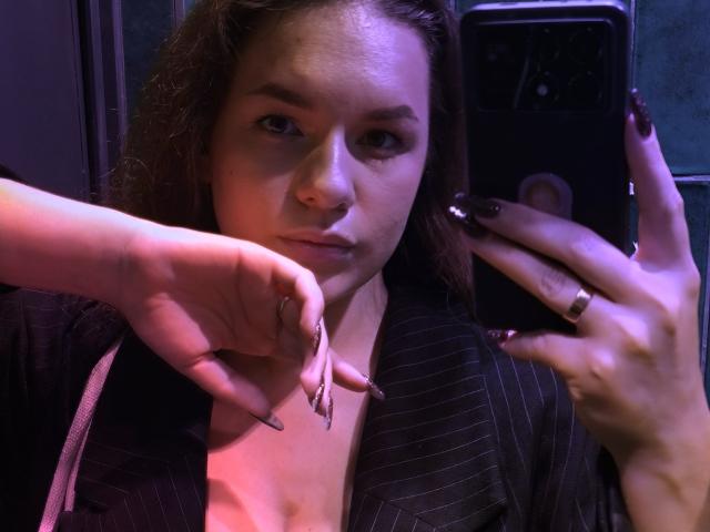 Dreia with brown hair and nose ring, wearing pinstriped shirt, taking selfie with purple-tinged light. Holding chain necklace with left hand, fingernails painted dark.