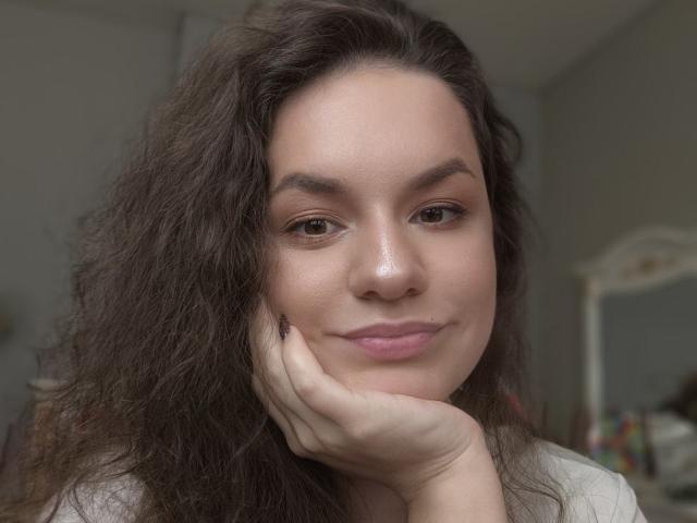 Dreia, met golvend bruin haar, leunt haar wang op haar hand en glimlacht zachtjes. Haar bruine ogen en roze lippen vallen op. Ze draagt een lichtgrijze blouse..