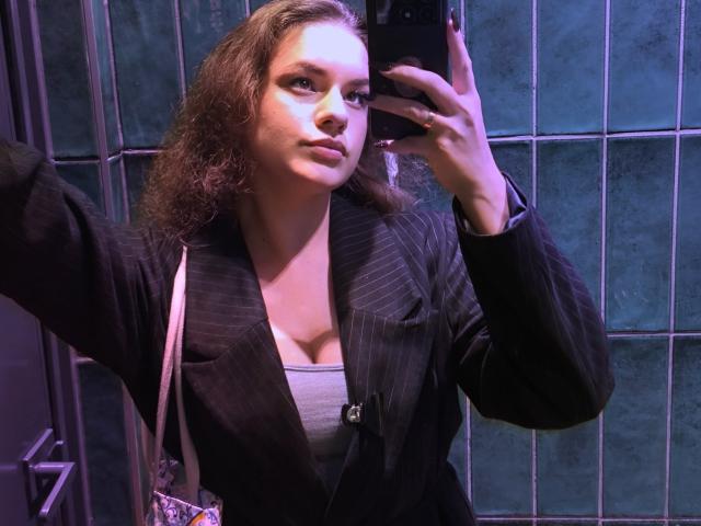 Dreia with curly brown hair, wearing a pinstriped black blazer over a gray tank top, taking a mirror selfie, showing her boobs, in a dimly lit room.